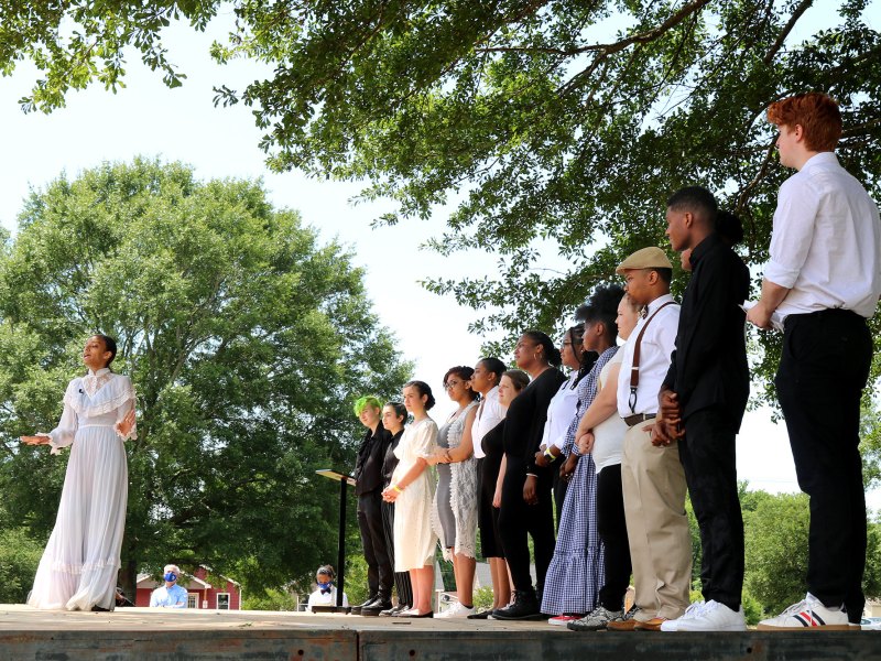 Students stand on a large outdoor stage in old fashioned clothes under large trees