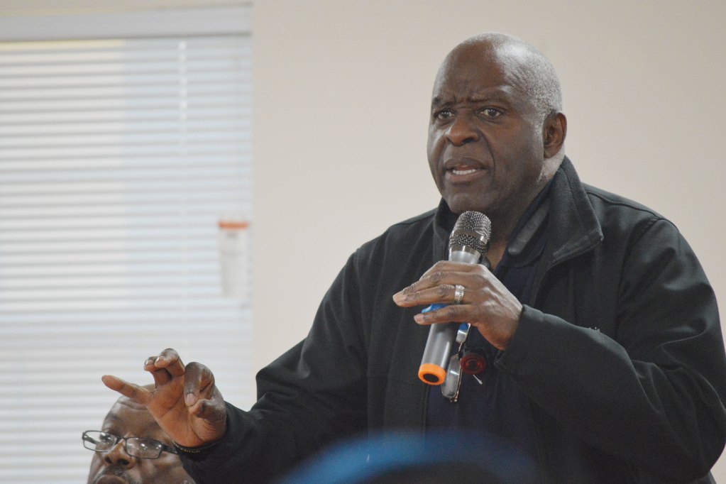 A man in a black jacket speaks while holding a grey mic