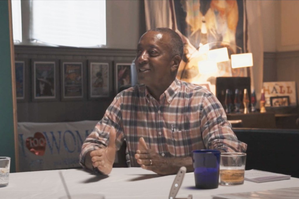 A man in a striped shirt sits at a table, gesturing with his hands as he speaks.