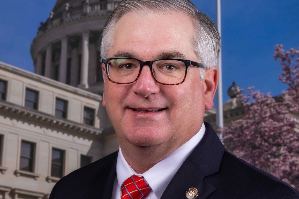 Official portrait of a man in glasses and a dark suit standing before the Mississippi state capital building