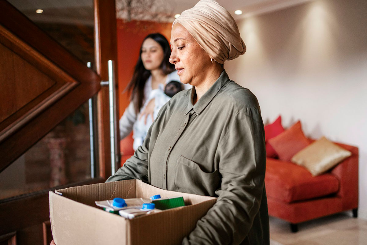 A woman in a head covering is carrying a cardboard box of items somewhere. A woman holding a baby is seen in the background of the living room
