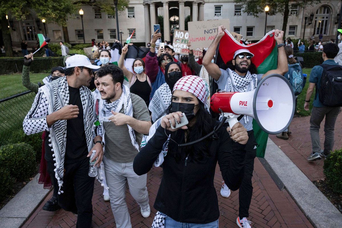 A woman with a red and white scarf on her head leads a protest of people.