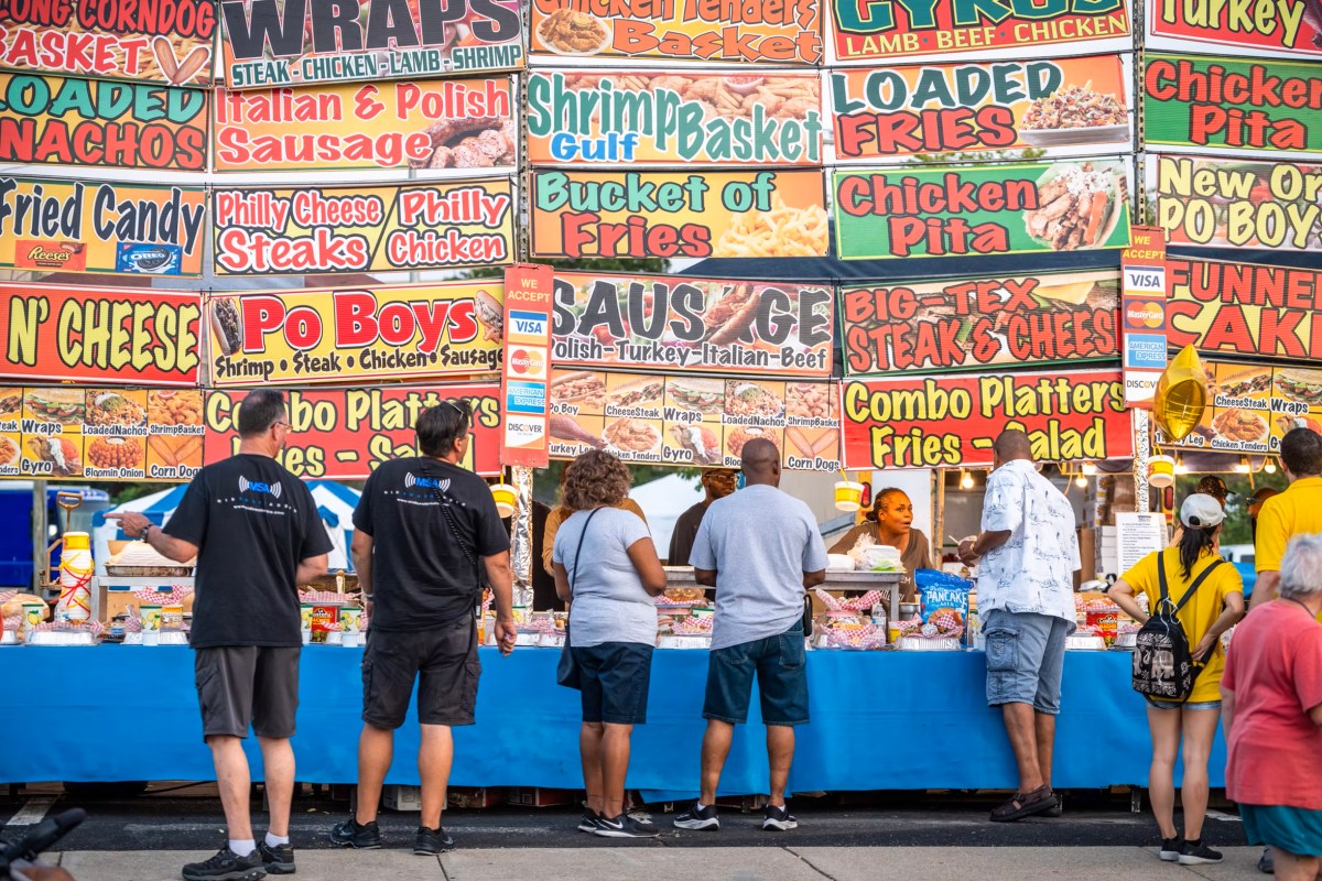 People stand in front of a fair food vendor with a multitude of giant colorful food signs