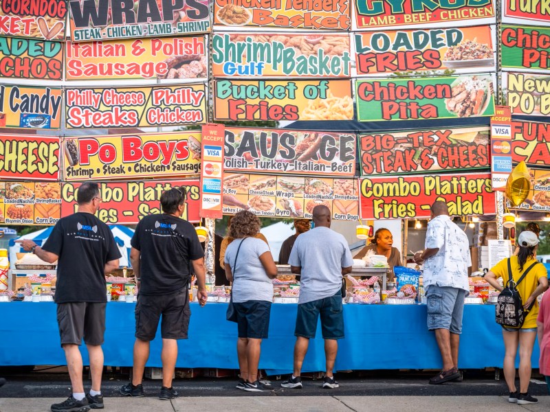 People stand in front of a fair food vendor with a multitude of giant colorful food signs