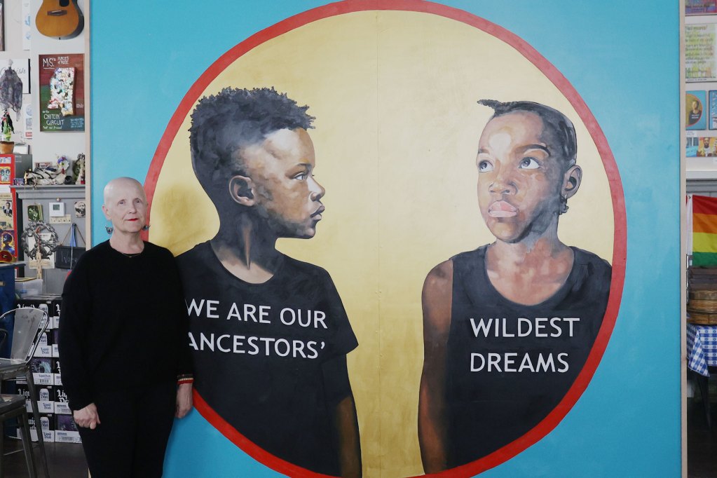A woman in black attire stands to the left of a large artwork depicting two boys whose shirts say, “We are our ancestors’ wildest dreams.”