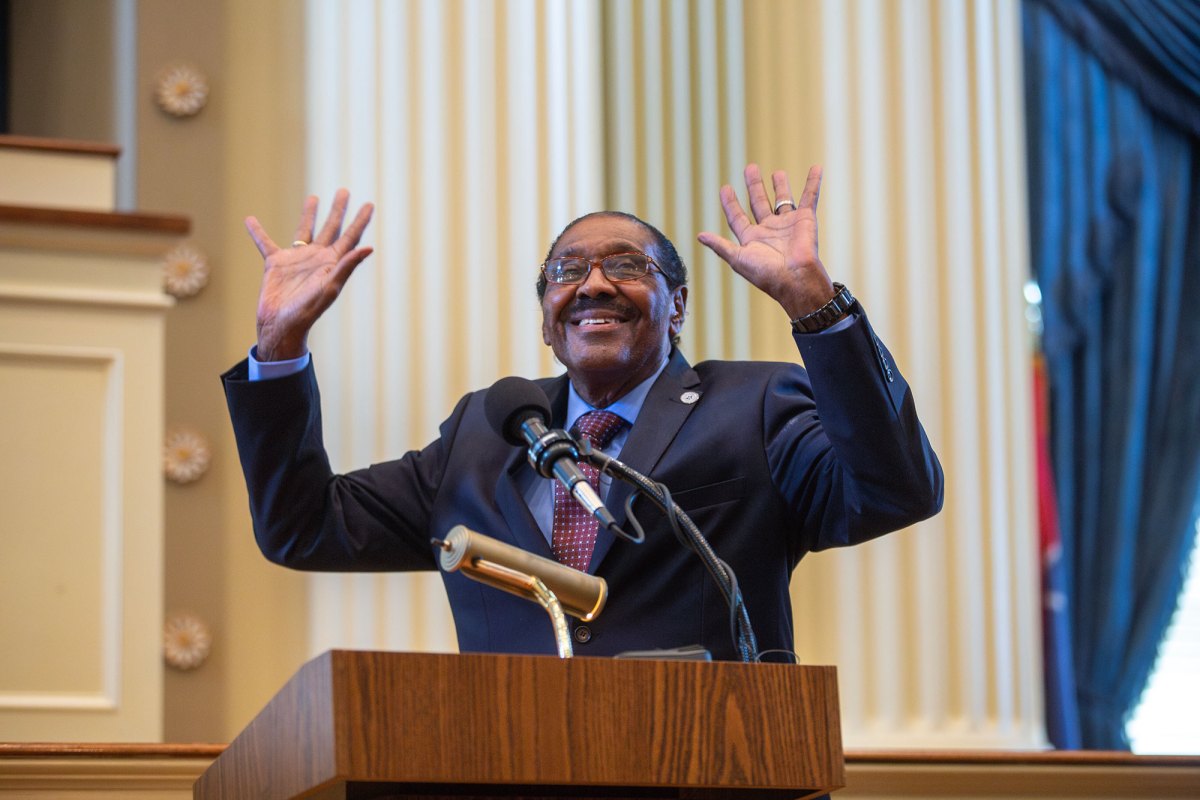 A man in a suit smiles and waves both hands from behind a wooden podium