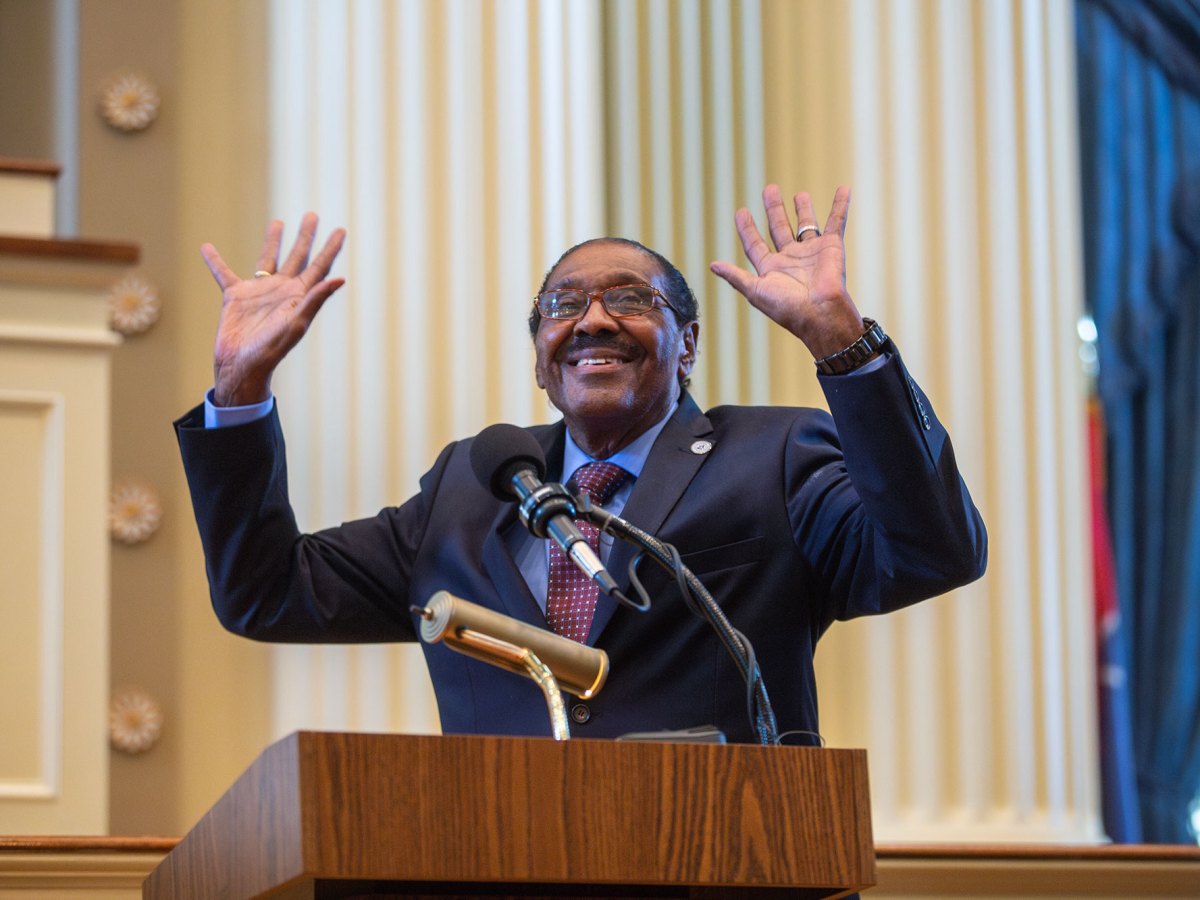 A man in a suit smiles and waves both hands from behind a wooden podium