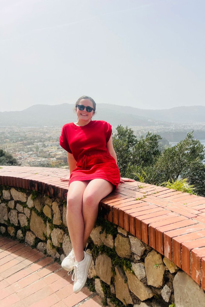 A young woman wearing a red dress and black sunglasses sits on a brick structure with a vast landscape behind her
