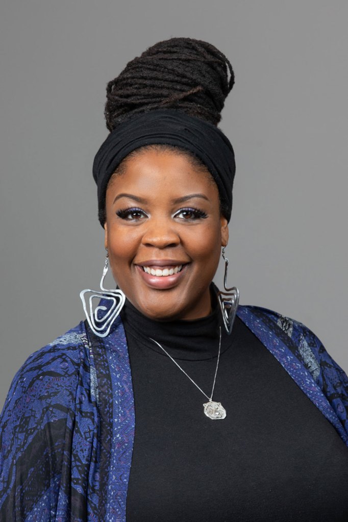 Headshot of a woman in black and blue clothes with large symmetrical silver earrings and her hair up in a large bun on top of her head