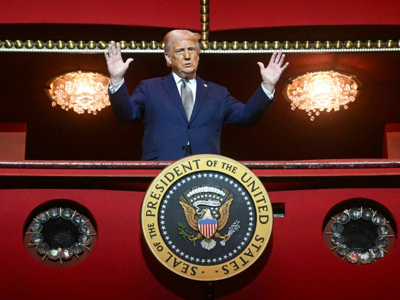 Donald Trump is standing at a red balcony holding his hands up and looking down. A presidential seal is affixed to the red balcony and two chandeliers glow behind him on a red cieling