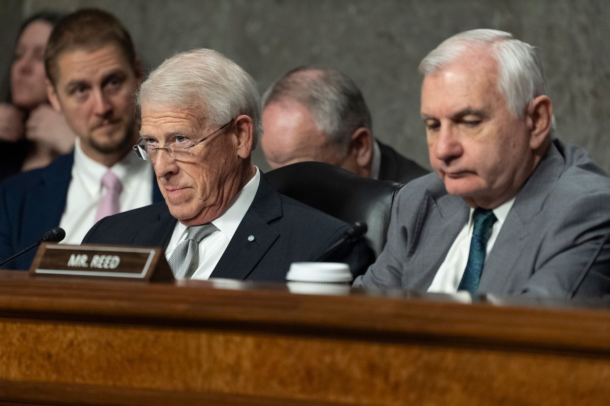 Sen. Roger Wicker and Sen. Jack Reed sit in a public meeting behind brown wood with a mr. reed name plate on top of it.