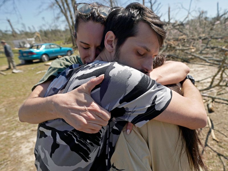 Three people embrace in the middle of a tornado damaged neighborhood