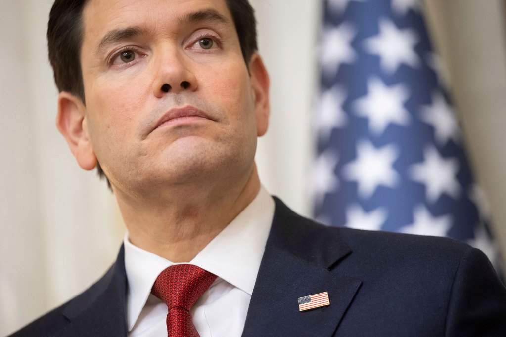 A man in suit with a red tie stands in front of an American flag.
