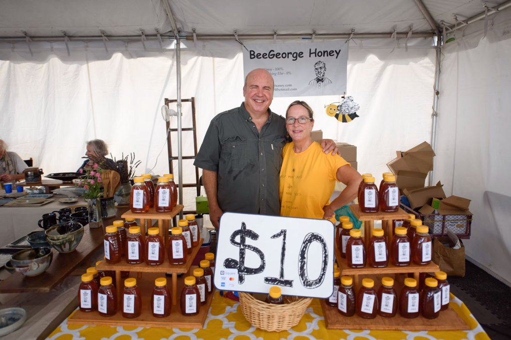 Two people stand at a vendor stand labeled BeeGeorge Honey, with a table display of $10 bottles of honey