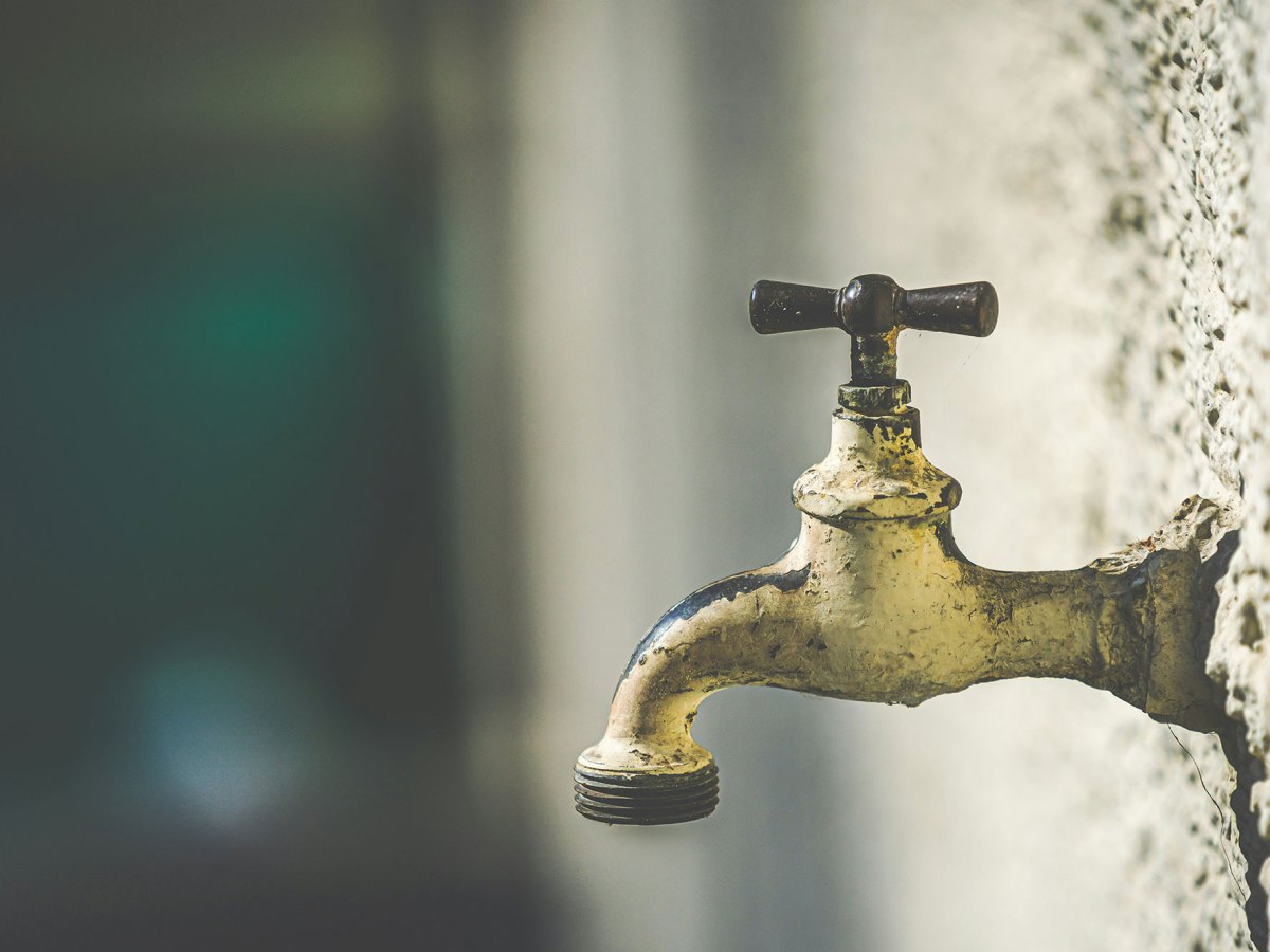 Pictured is a white rusty water faucet coming out of the side of a popcorn textured wall.