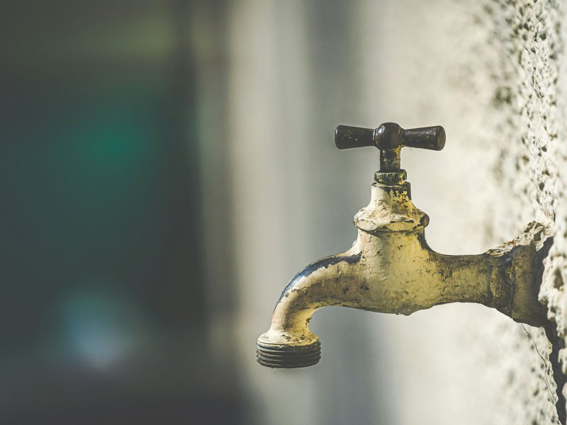 Pictured is a white rusty water faucet coming out of the side of a popcorn textured wall.