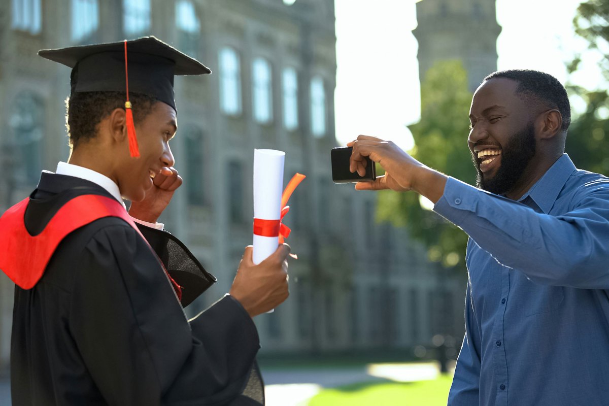 Young man in black cap and gown smiling at man holding camera in blue shirt