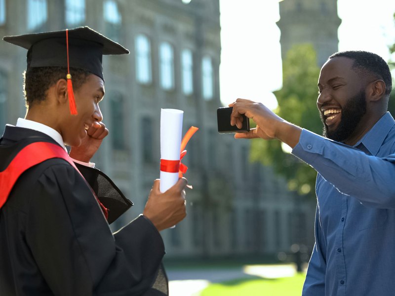 Young man in black cap and gown smiling at man holding camera in blue shirt