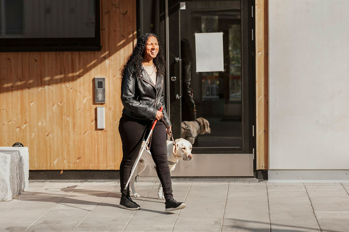 A woman in a moto jacket walks on a sidewalk with a walking cane in one hand and a seeing eye dog in the other