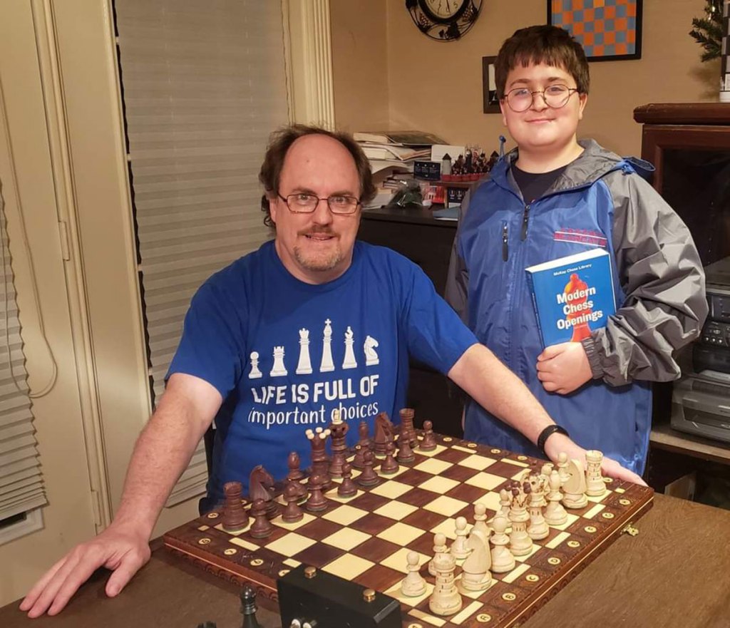 A man sits at a table with a chess board set up before him. A teen stands beside him holding a book labeled Master Chess Openings
