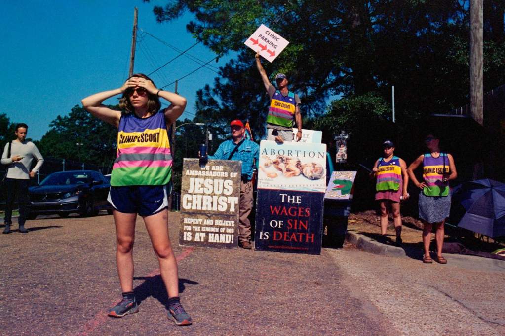 A clinic escort holds her hands over her forehead, looking in the distance, while anti-abortion protesters with grotesque signs depicting dead fetuses standing behind her, one saying "We Are Ambassadors For Jesus Christ." Behind the protesters, a man waves a sign that says "Clinic Entrance" with an arrow pointing right