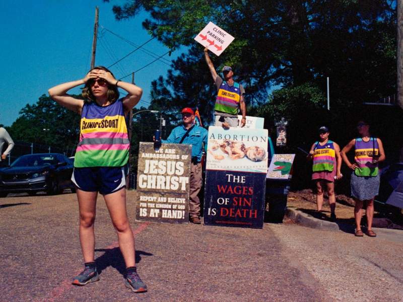 A clinic escort holds her hands over her forehead, looking in the distance, while anti-abortion protesters with grotesque signs depicting dead fetuses standing behind her, one saying "We Are Ambassadors For Jesus Christ." Behind the protesters, a man waves a sign that says "Clinic Entrance" with an arrow pointing right
