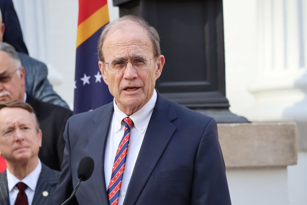 Lieutenant Governor Delbert Hosemann speaking at a presser with a red and blue tie and the Mississippi state flag behind him