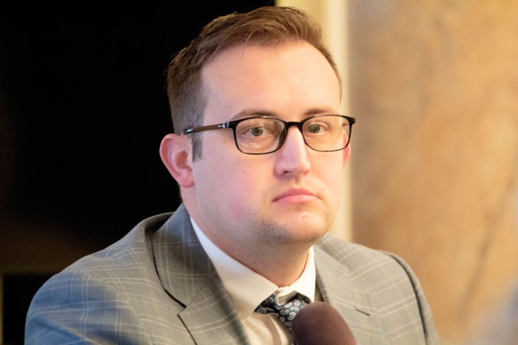 A man sits in front of a mic wearing a grey plaid shirt and black glasses