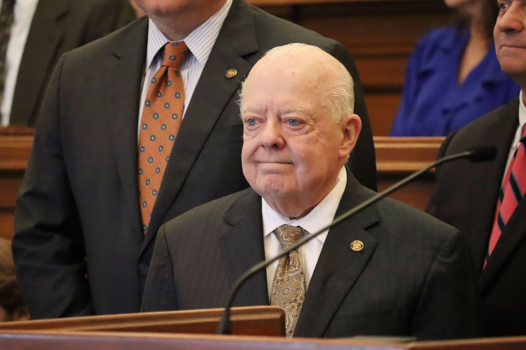 Man in black suit and white shirt with a yellow tie stands near a microphone with tears in his eyes