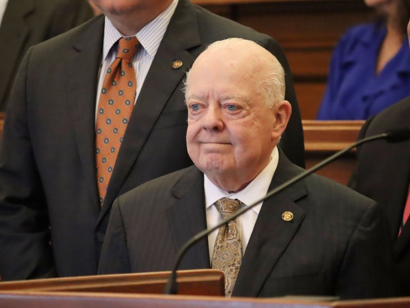 Man in black suit and white shirt with a yellow tie stands near a microphone with tears in his eyes