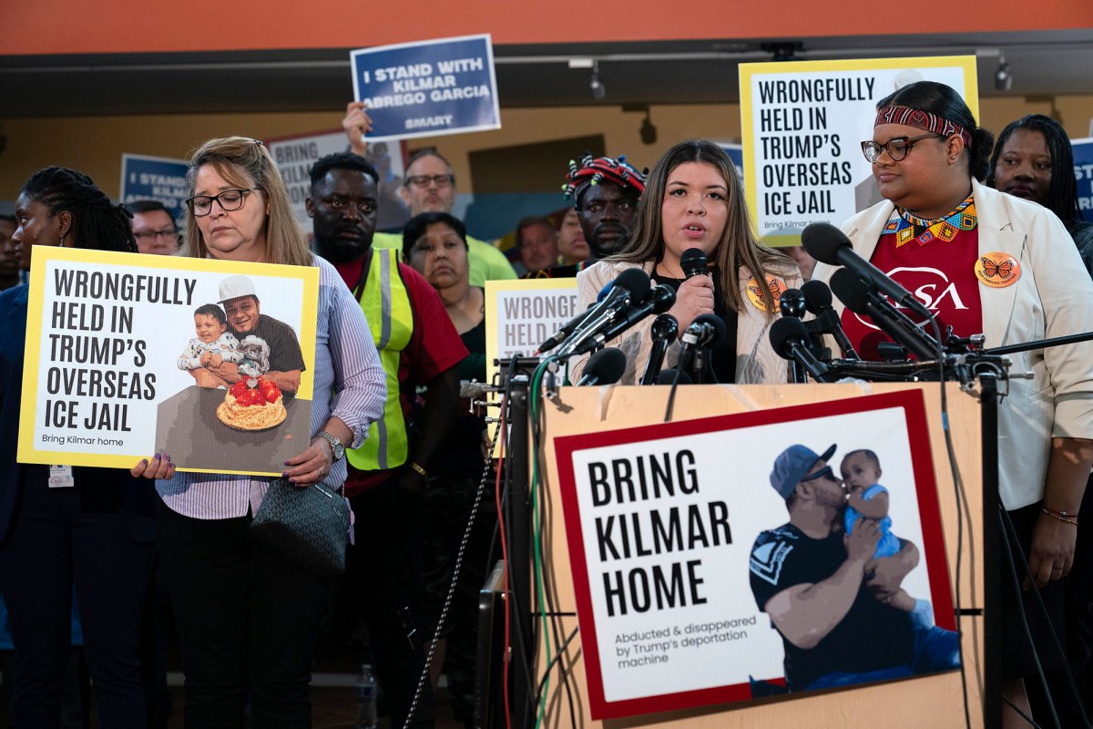 A woman speaking at a podium with mics is surrounded by people holding white, yellow, and blue signs