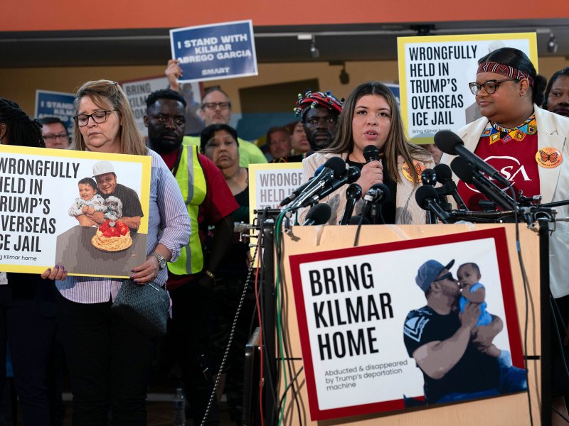 A woman speaking at a podium with mics is surrounded by people holding white, yellow, and blue signs