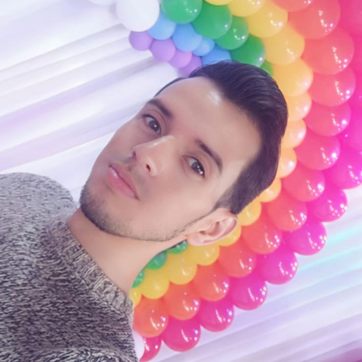 A clean-cut man with brown hair and brown eyes looking at the viewer while he stands in front of rainbow balloons