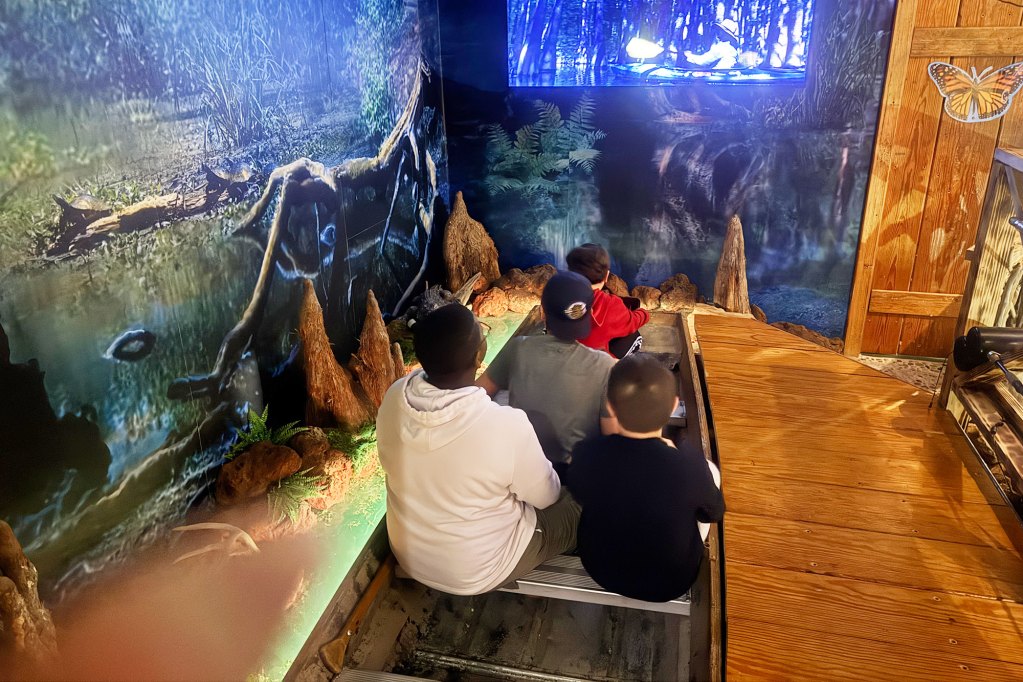 Four boys sit inside of a boat, while gazing at a projected image on the wall. The room also features swamp wallpaper