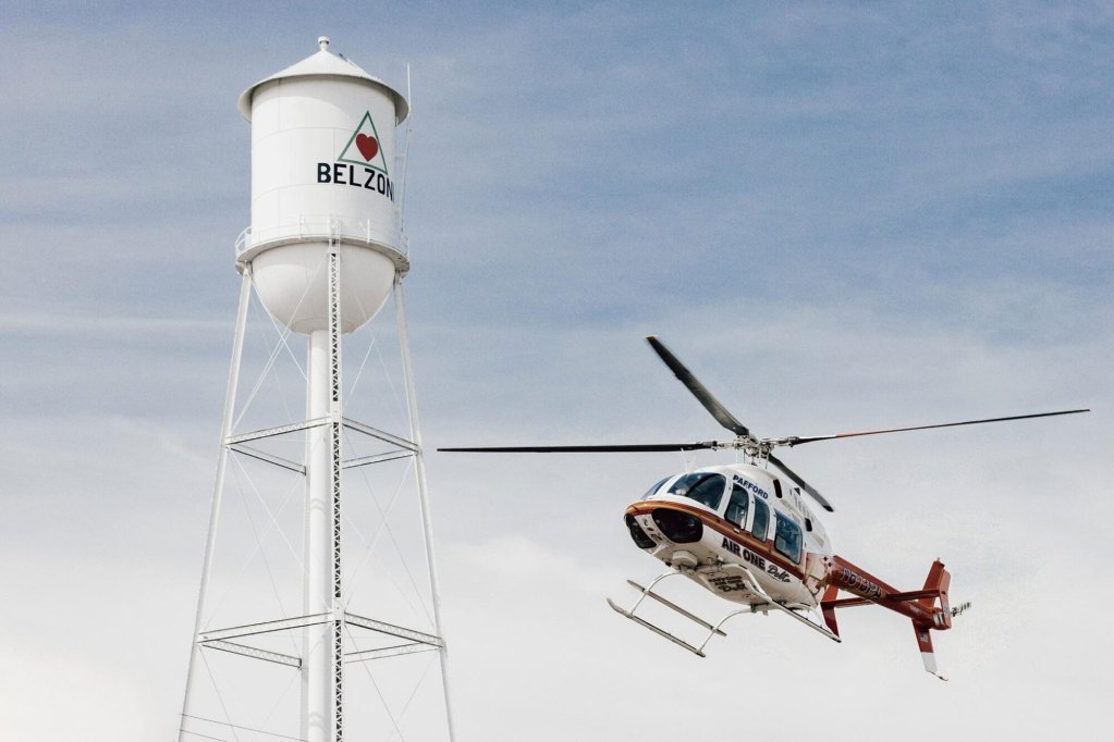 A white and maroon helicopter flies right next to a tall white water tower with Belzoni written on it.