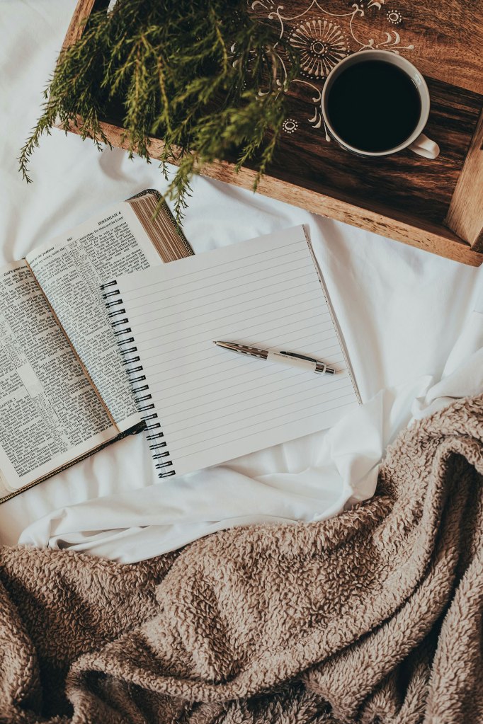 A warm photo of a bible, notepad, and pen on a bed with white sheets; surrounded by a brown blanket and a wooden tray with a cup of coffee.
