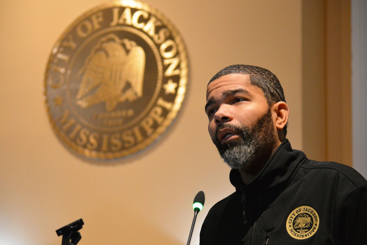 Chokwe A Lumumba seen in a back jacket with seal of the City of Jackson Mississippi on the chest speaks at a mic. The seal is seen again on the wall behind him.
