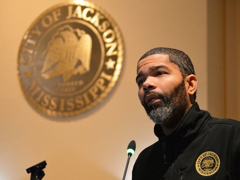 Chokwe A Lumumba seen in a back jacket with seal of the City of Jackson Mississippi on the chest speaks at a mic. The seal is seen again on the wall behind him.