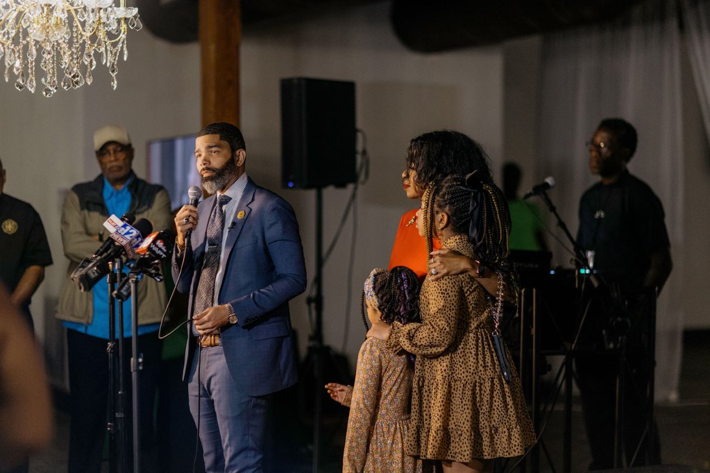 a photo of a man in a suit speaking on a stage with other people standing around him, including a woman and two young girls