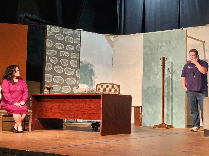 Two people perform on a theater stage set up like an office
