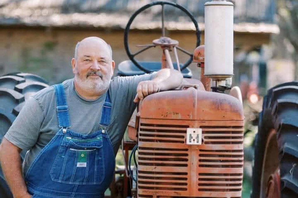 A man in blue overalls standing next to a rusty orange tractor