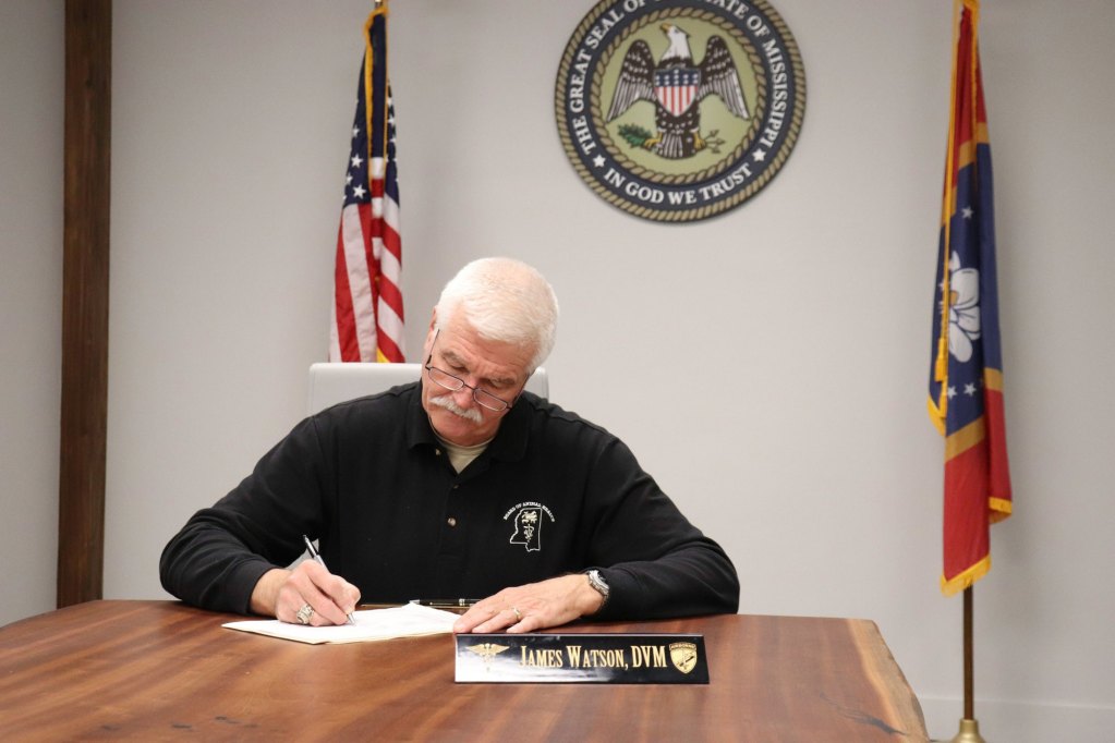 Man in black official shirt signing documents sitting at a wooden table with the American flag, state flag, and the great seal of the state of Mississippi behind him