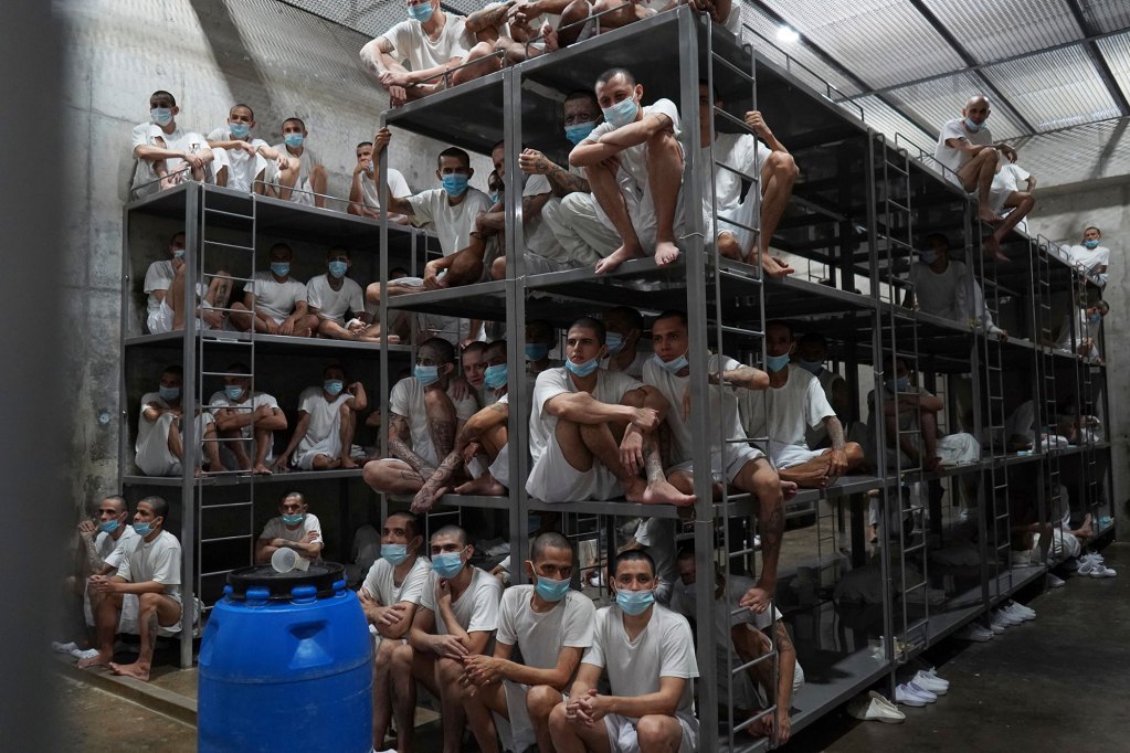 A view inside a large population jail cell with rows of four level bunk beds filled with masked inmates in white underclothes