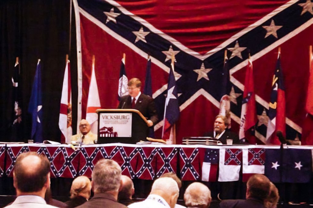 Tate Reeves speaking at a podium surrounded by Confederate flags.