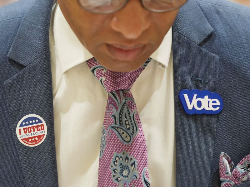 Closeup of a person in a suit's chest, showing an I Voted in Hinds County sticker on the left lapel, and a Vote pin in the right.