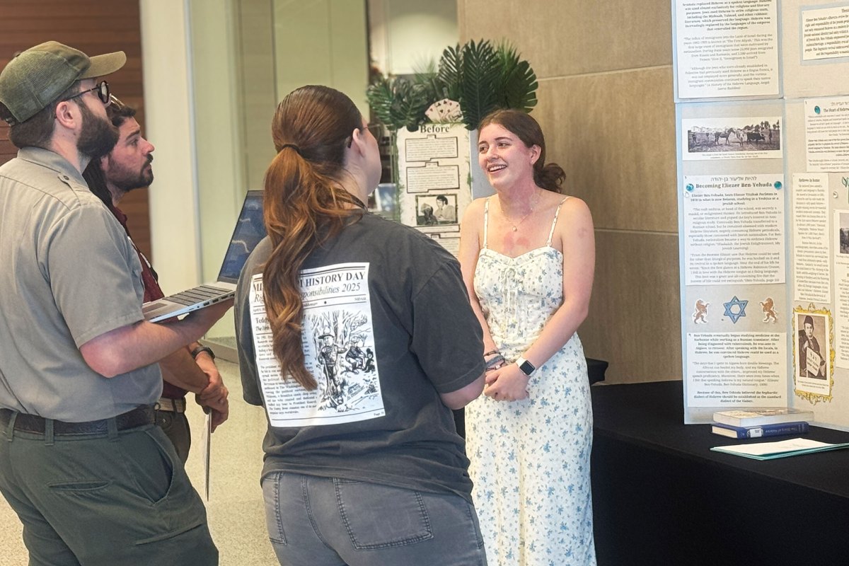 A young woman in a white dress explains her research to a trio of judges.