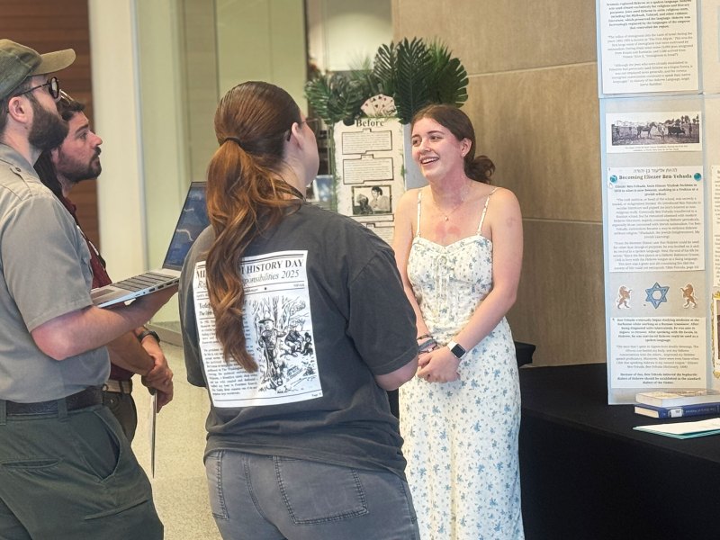 A young woman in a white dress explains her research to a trio of judges.