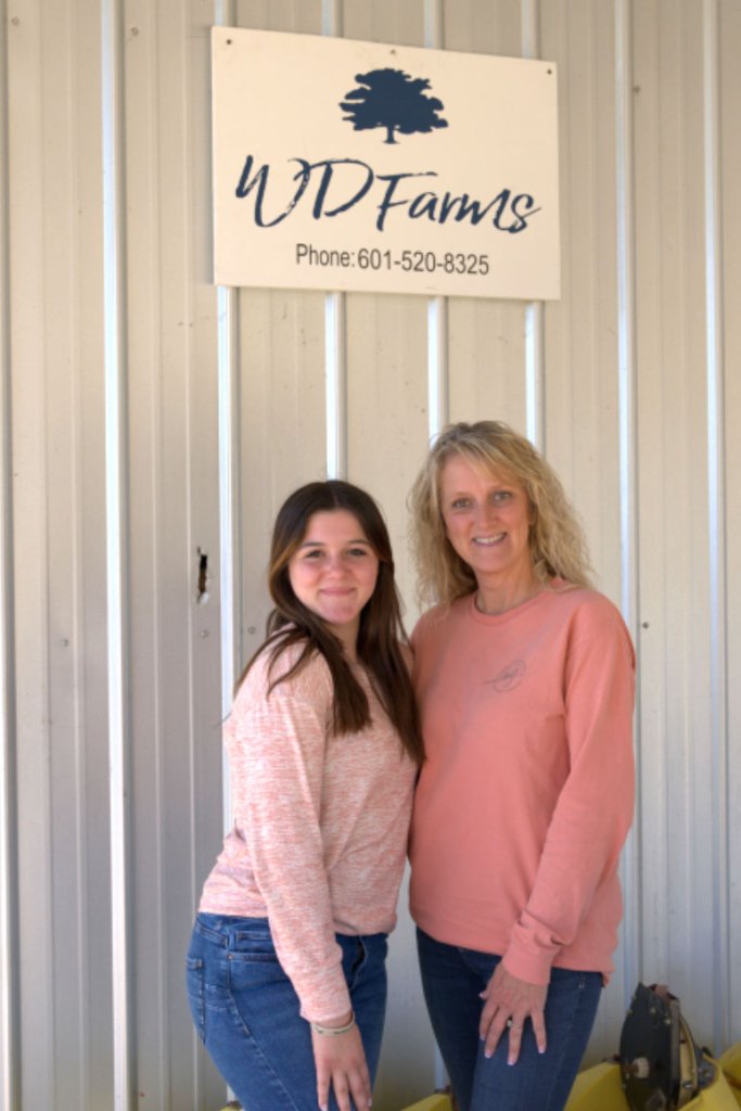 Two women pose in front of a metal building with a sign that reads WD Farms