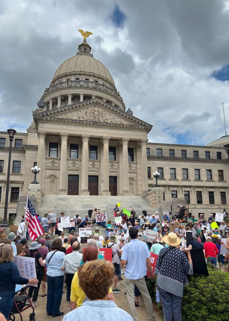 a photo shows the Mississippi Capitol with a gold eagle atop its dome, and dozens of people gathered at its steps holding protests signs that say things like, "Tax the Rich, Fire Musk" and "Impeach Trump, Fire Elon." One person is holding an American flag