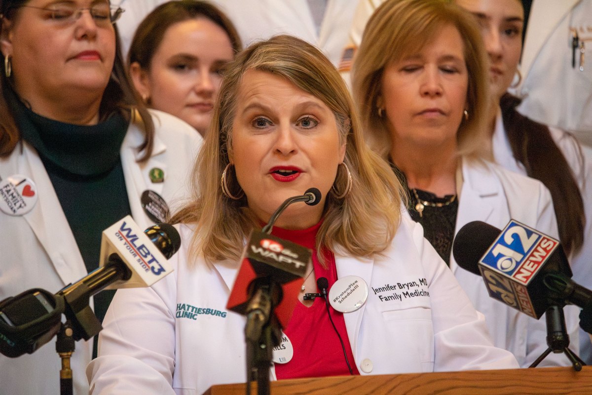 An woman in a doctor's coat speaks at a podium, other women in lap coats behind her